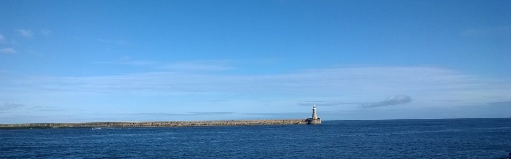 Vista looking out to see with light blue sky, deep blue sea and a lighthouse at the end of a pier