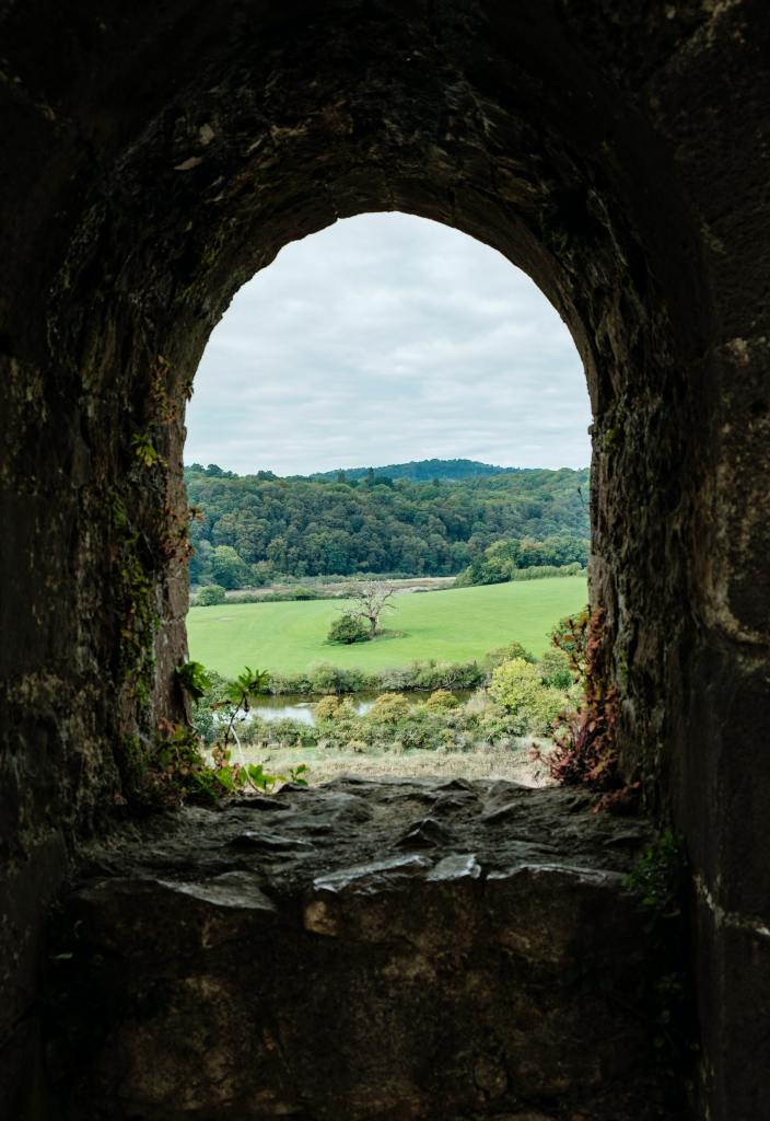 
Stone archway leading the eye over to a countryside scene, with a river in the foreground, a tree in a field in the midground and a hill in the background.